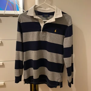 Rugbytröja från Polo Ralph Lauren - Stilren och klassisk rugbytröja (The Iconic Rugby Shirt) från Polo Ralph Lauren i marinblått och grått. Storlek 10-12 år eller M. Tröjan är använd men är i mycket fint skick. Listpris 1195 kr, säljes för 550 kr.