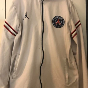 Vit jacka från Jordan med PSG-logga - Säljer en vit jacka från Jordan med PSG-logga på bröstet. Jackan har dragkedja och långa ärmar med röda och svarta ränder. Perfekt för sportiga tillfällen eller som en stilren vardagsjacka.