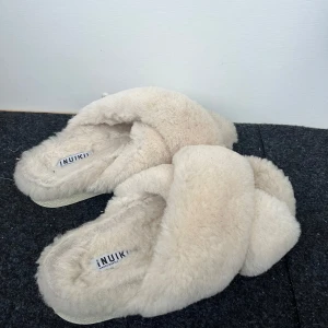 Beige fluffiga tofflor från Inuikii - Mysiga och varma beige tofflor från Inuikii med korsade remmar och en mjuk, fluffig yta. Perfekta för att hålla fötterna varma under kyliga dagar. Sulan är robust och de är i bra skick, använda men har massor kvar att ge till nästa ägare😇💓 