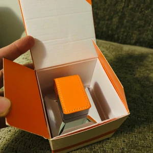 Saga Eau de Parfum från Emper - Upptäck Salga Eau de Parfum från Emper, en elegant doft i en stilren flaska. Flaskan har en fyrkantig form med en silverfärgad topp och ett distinkt orange lock. Förpackningen matchar med en snygg design i vitt och orange.