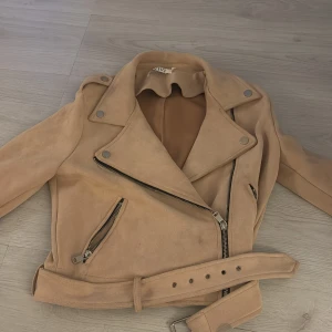 Beige mockajacka från IVIVI - Snygg beige mockajacka från IVIVI med dragkedja och bälte. Jackan har en klassisk bikerstil med detaljer som knappar och snedställda fickor. Perfekt för en trendig look. Pris kan diskuteras