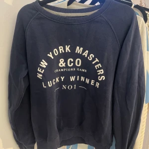 Mörkblå sweatshirt med tryck - Säljer en mörkblå sweatshirt med texten 'New York Masters & Co Lucky Winner No1' i vitt. Tröjan har långa ärmar och en rund halsringning. Perfekt för en avslappnad stil.