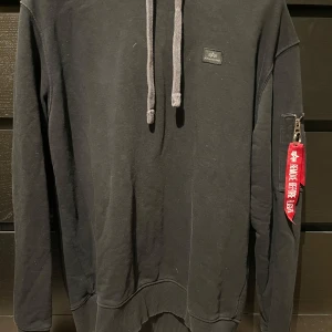 Svart hoodie från Alpha Industries - Säljer en svart hoodie från Alpha Industries med en cool 'Remove Before Flight'-detalj på ärmen. Hoodien har en ficka med dragkedja på ärmen och justerbara snören i huvan. Insidan är i en kontrasterande orange färg. Produkten är inköpt i fysisk butik på Väla i Helsingborg på Carlings. Knappt använd. 
