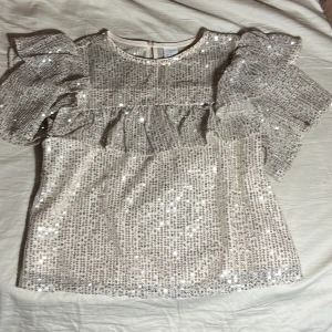 Glittrig topp från Lindex Kids - Säljer en glittrig topp från Lindex Kids i silver med volangdetaljer. Toppen har korta ärmar och en knapp i nacken. Perfekt för festliga tillfällen! ✨