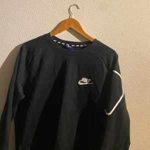 Svart tröja från Nike - Säljer en svart tröja från Nike med vit logga på bröstet och en dragkedja på ärmen. Tröjan har långa ärmar och en vit randdetalj längs ärmen. Perfekt för en sportig look!
