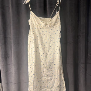 Vit blommig klänning från NA-KD - vit klänning från NA-KD med blått blommönster. Klänningen har smala axelband och knytning framtill. Slits på sidan.