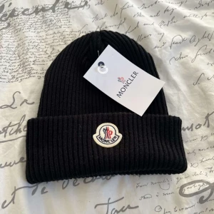 Svart ribbad mössa från Moncler - Snygg svart ribbad mössa från Moncler med broderad logotyp framtill. Perfekt för kyliga dagar och ger en stilren look. Tillverkad i mjukt material för extra komfort.