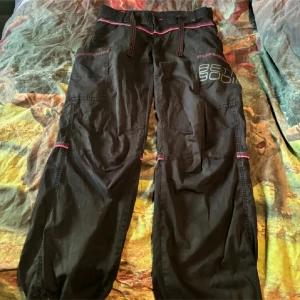 Svarta cargobyxor med rosa detaljer - Snygga svarta cargobyxor med rosa sömmar och snörning i midjan. Byxorna har flera fickor och en avslappnad passform, perfekt för en cool och bekväm stil.