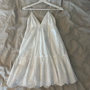 Vit sommarklänning med spetsdetaljer - Snygg vit klänning med smala axelband och v-ringning. Klänningen har vackra spetsdetaljer och en volangkant som ger en romantisk känsla. Perfekt för varma sommardagar. Eller skolavslutning. Slutsåld i butik