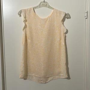 Gulmönstrad blus med volangdetaljer - Säljer en söt blus med gult mönster och volangdetaljer. Blusen är ärmlös och har en knäppning bak. Perfekt för en somrig look!