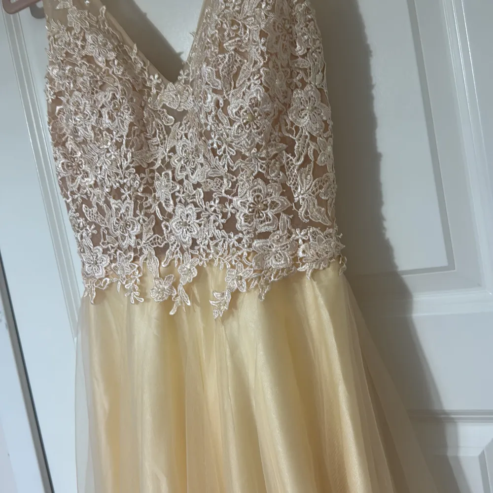 Elegant långklänning i champagnefärg med v-ringad spetsöverdel och blommönster. Kjolen är i ett lätt och flödande material som ger en vacker siluett.. Mekot.