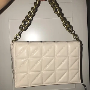 Beige quiltad axelväska med kedja - Snygg beige axelväska med quiltat mönster och en kraftig guldfärgad kedja som handtag. Väskan har en avtagbar axelrem för extra flexibilitet. Perfekt för att ge en stilren touch till din outfit.