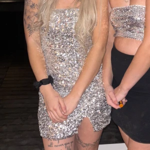 Glittrig silverklänning - Snygg och glittrig silverklänning med paljetter. Klänningen är kort och axelbandslös, perfekt för en festlig kväll. Den har en tight passform som framhäver figuren.