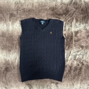  Polo Ralph Lauren - Säljer en snygg mörkblå stickad väst från Polo Ralph Lauren. Västen har en klassisk v-ringning och ett diskret broderat märke på bröstet. Perfekt för lager-på-lager under kyligare dagar.