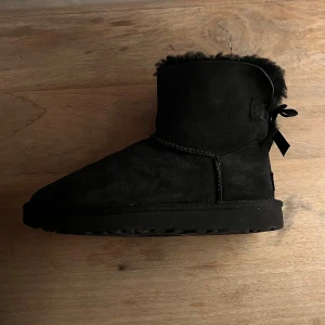 UGG - Svarta boots med rosett ur fluff   - Snygga och bekväma svarta boots från UGG med rosett-detaljer på baksidan. Tillverkade i mocka med en mjuk pälsfodring för extra värme. Perfekta för kyliga dagar🥰🥰❤️❤️