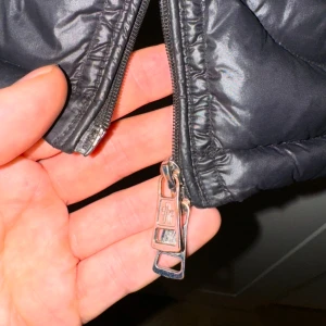 Svart dunjacka från Moncler - Snygg svart dunjacka från Moncler med quiltad design och dragkedja. Jackan har en klassisk look med långärmade ärmar och Moncler-logga på ärmen. Perfekt för kyligare väder.