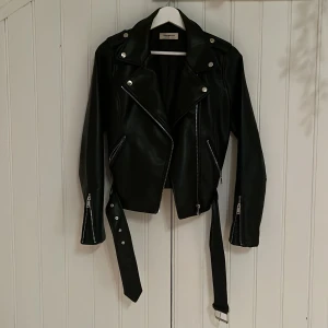 Svart skinnjacka från Nelly - Snygg svart skinnjacka från Nelly med dragkedjor och bälte. Jackan har en klassisk bikerstil med långa ärmar och silverfärgade detaljer. Perfekt för en edgy look!