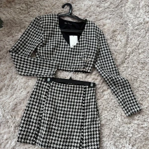 Svartvit rutig croppad topp från Zara + kjol - Snygg croppad topp från Zara i svartvitt hundtandsmönster i tweet material. Toppen har en v-ringad design med långa ärmar och en elegant krokdetalj framtill. Perfekt för en stilren look. Helt ny oanvänd saknas en mini diamant men inget som syns 
