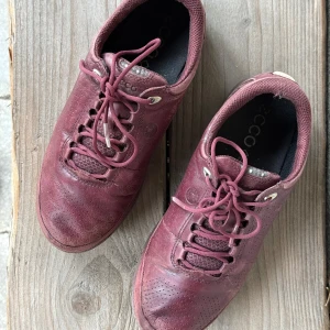 Vinröda sneakers från Ecco - Gore-Tex vinröda sneakers från Ecco med snörning och perforerade detaljer. Skorna har en rund tå och är tillverkade i skinn, vilket ger en stilren look. Perfekta för en avslappnad stil.