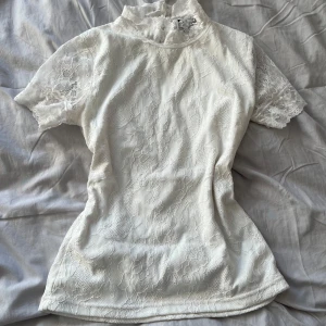 Vit spetsblus - Vit spetsblus med korta ärmar och hög krage. Blusen har ett vackert blommönster och en knappdetalj i nacken. Passar som XS/S.