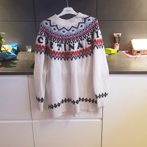 Vit stickad tröja från H&M - Säljer en vit stickad tröja från H&M med ett färgglatt mönster i svart, rött och blått. Tröjan har långa ärmar och ett ribbat mönster vid ärmslut och nederkant. Perfekt för kyliga dagar!