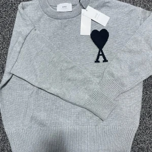 Grå tröja från Ami - Snygg grå tröja från Ami med ett svart hjärtmönster och bokstaven A på bröstet. Tröjan har ribbade muddar och rund halsringning. Perfekt för en stilren look.