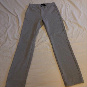 Grå kostymbyxor från ELVINE - Snygga grå kostymbyxor med en klassisk straight passform. De har en diskret dragkedja och knapp framtill samt fickor bak med knappdetalj. Perfekta för en stilren look.