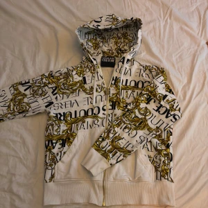 Vit hoodie från Versace Jeans Couture - Snygg vit hoodie med ett iögonfallande mönster i svart och guld från Versace Jeans Couture. Den har en dragkedja framtill och en praktisk huva. Perfekt för att ge din outfit en lyxig touch.