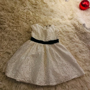 Vit spetsklänning från Lindex Kids. Storlek 98.  - Charmig vit spetsklänning från Lindex för barn. Klänningen har en söt svart rosett i midjan och korta ärmar. Perfekt för speciella tillfällen.