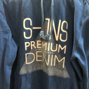 Blå hoodie med tryck - Snygg blå hoodie med texten 'S-INS PREMIUM DENIM' tryckt på framsidan. Den har en justerbar dragsko i huvan och en klassisk känguruficka. Perfekt för en avslappnad stil.