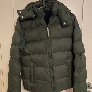 Grön dunjacka från GANT - Säljer en snygg grön dunjacka från GANT storlek XS. Jackan har en quiltad design och är perfekt för kyliga dagar. Den har en avtagbar huva och broderad logotyp på bröstet. Bra pris pga ett skrapmärke på ryggen men förutom det så är den i perfekt skick.