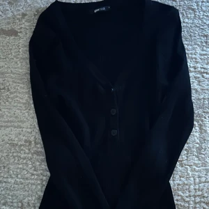 Svart kofta från Gina Tricot - Säljer en stilren svart kofta från Gina Tricot i storlek S. Koftan har en v-ringad design med knappar framtill och långa ärmar. Perfekt för lager-på-lager-stil.