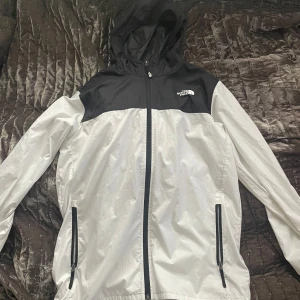Svart och vit vindjacka från The North Face - Säljer en snygg och praktisk vindjacka från The North Face i storlek s. Jackan är svart och vit med dragkedja framtill och två fickor med dragkedjor. Perfekt för vår och höst med sin lätta och bekväma design. Igentligen är den junior xl passform S