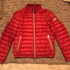 Säljer en snygg röd dunjacka från Moncler. Jackan är i nyskick och har en stilren design med dragkedja och Monclers logga på ärmen. Perfekt för höst och vinter, håller dig varm och trendig. Den har en normal passform och är långärmad. Passar perfekt för både vardag och fest!