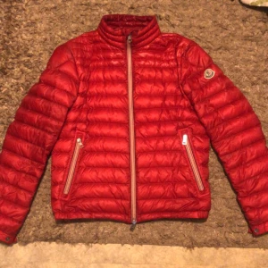 Röd dunjacka från Moncler - Säljer en snygg röd dunjacka från Moncler. Jackan är i nyskick och har en stilren design med dragkedja och Monclers logga på ärmen. Perfekt för höst och vinter, håller dig varm och trendig. Den har en normal passform och är långärmad. Passar perfekt för både vardag och fest!