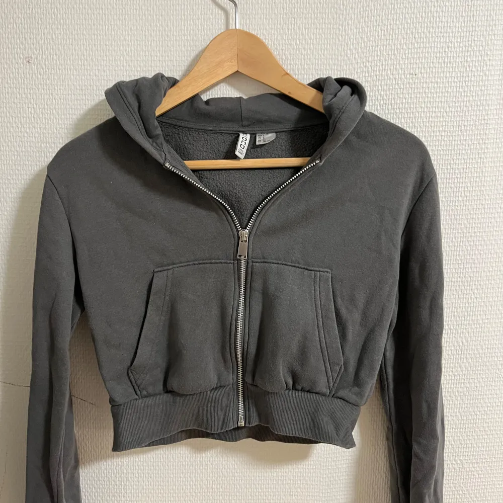 Säljer en supermjuk och bekväm grå hoodie från H&M. Den är croppad och har en dragkedja framtill, perfekt för en avslappnad look. Hoodien har långa ärmar och en mysig huva. Perfekt för höst och vår!. Tröjor & Koftor.