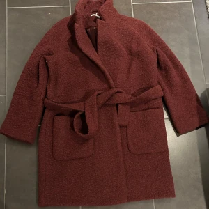 Ganni ullkappa - En lång ullkappa i bra skick, vinröd. Den har en krage, en knäppning framtill och en bälte i midjan. Den har två fickor på framsidan. Jag är 162 cm och kappan slutar precis ovanför knäna. Nypris ca 3500:-