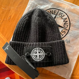 Svart ribbad mössa från Stone Island - Säljer en snygg svart ribbad mössa från Stone Island. Den har en cool broderad logga framtill och är perfekt för att hålla sig varm under höst och vinter. Mössan är i nyskick och kommer med originalförpackning. Passar perfekt för både vardag och lite mer uppklädda tillfällen.