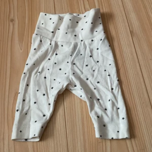 Vita byxor med stjärnmönster från H&M - Säljer ett par supersöta vita byxor från H&M med små svarta stjärnor över hela tyget. De är gjorda av ekologisk bomull, vilket gör dem mjuka och sköna för de allra minsta. Perfekta för bebisar i åldern 0-1 månad. Byxorna har en bekväm passform och är lätta att ta på och av. Ett måste för den lilla stjärnan i ditt liv! 🌟