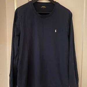 Mörkblå långärmad tröja från Ralph Lauren - Säljer en stilren och klassisk mörkblå långärmad tshirt från Ralph Lauren. Tröjan har en liten vit logga på bröstet och är perfekt för både vardag och lite finare tillfällen. Den är i bra skick och superbekväm att ha på sig. Passar perfekt till höst.