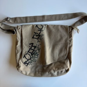 Beige axelväska med tryck - Säljer en beige axelväska i slitstarkt material med ett tryck på framsidan. Väskan har en justerbar axelrem och ett stort huvudfack som stängs med ett lock.m!