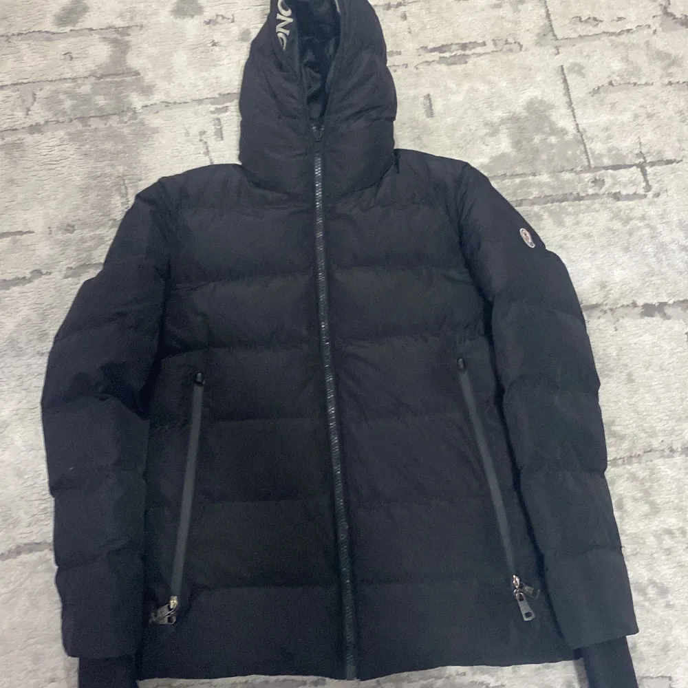 Säljer en snygg och varm svart dunjacka från Moncler. Jackan har en stilren design med dragkedja och Moncler-logga på ärmen. Perfekt för kalla vinterdagar. Den är i mycket braskick passform. Huvan är justerbar och insidan har Moncler-tryck l. Jackor.