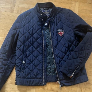 Mörkblå quiltad jacka från Morris - Säljer en snygg mörkblå quiltad jacka från Morris. Den har en cool broderad patch på bröstet och stängs med dragkedja och knappar. Jackan är perfekt för höst och vår, med en stilren design och bekvämt foder. Passar både till vardags och fest!