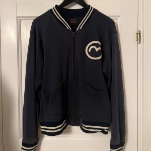 Mörkblå bomberjacka från Evisu - Säljer en snygg mörkblå bomberjacka från Evisu. Den har vita ränder på kragen, ärmsluten och nederkanten. På framsidan finns en vit logga och på baksidan står det 'MIGHTY EVISU' i stora bokstäver. Fixad nu zipper, strl L men passar m/s