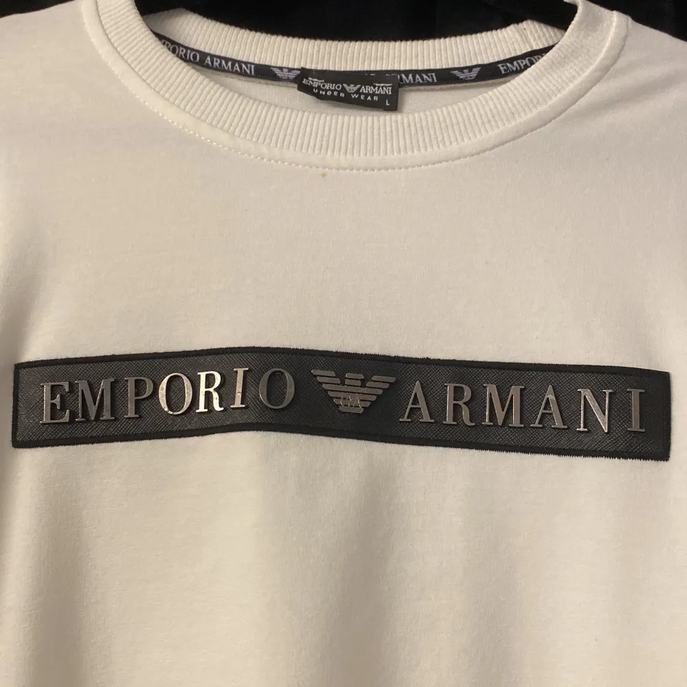 Säljer en stilren vit tröja från Emporio Armani. Den har en klassisk design med ett svart märke på bröstet där det står 'Emporio Armani'. Tröjan är långärmad och har en normal passform, perfekt för både vardag och lite finare tillfällen. Materialet känns mjukt och bekvämt. Perfekt för dig som vill ha något enkelt men ändå snyggt i garderoben!. Tröjor & Koftor.