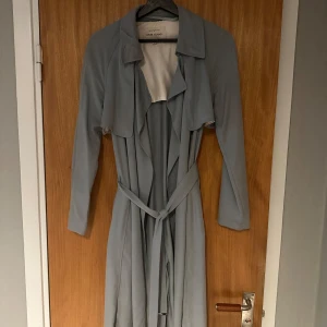 Ljusblå trenchcoat från River Island - Säljer en snygg ljusblå trenchcoat från River Island. Den har en klassisk design med ett bälte i midjan och långa ärmar. Perfekt för vår och höst när du vill ha en stilren look. Jackan är i bra skick och redo för nya äventyr!