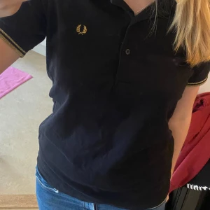 Pikétröja  - Säljer en svart Fred Perry t-shirt med guldiga detaljer på armarna och kragen. Väldigt bra skick endast använd fåtal gånger. Står stolek 44 men passar mer som en M/38. Jag brukar ha S/M i toppar💗☺️