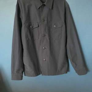 Grå overshirt med knappar - Säljer en stilren grå overshirt med knappar framtill. Den har två bröstfickor med knappar och är perfekt för lager-på-lager-stil. Passar både till vardags och lite mer uppklädda tillfällen.