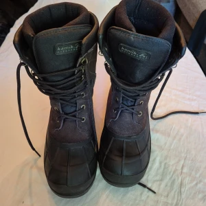 Svarta vinterkängor från Kamik - Säljer ett par robusta och varma vinterkängor från Kamik. De är svarta och har en hög design med snörning framtill. Kängorna är tillverkade i ett slitstarkt material som tål tuffa väderförhållanden. Sulan är grovmönstrad för extra grepp på hala underlag. Perfekta för vinterns kalla dagar!