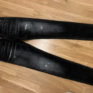 Svarta slitna jeans från Dsquared2 - Säljer ett par svarta jeans från Dsquared2 i storlek 44. De är snyggt slitna med flera hål och slitningar på både fram- och baksidan. Perfekta för en cool och avslappnad stil. De är i bra skick och redo för en ny ägare!